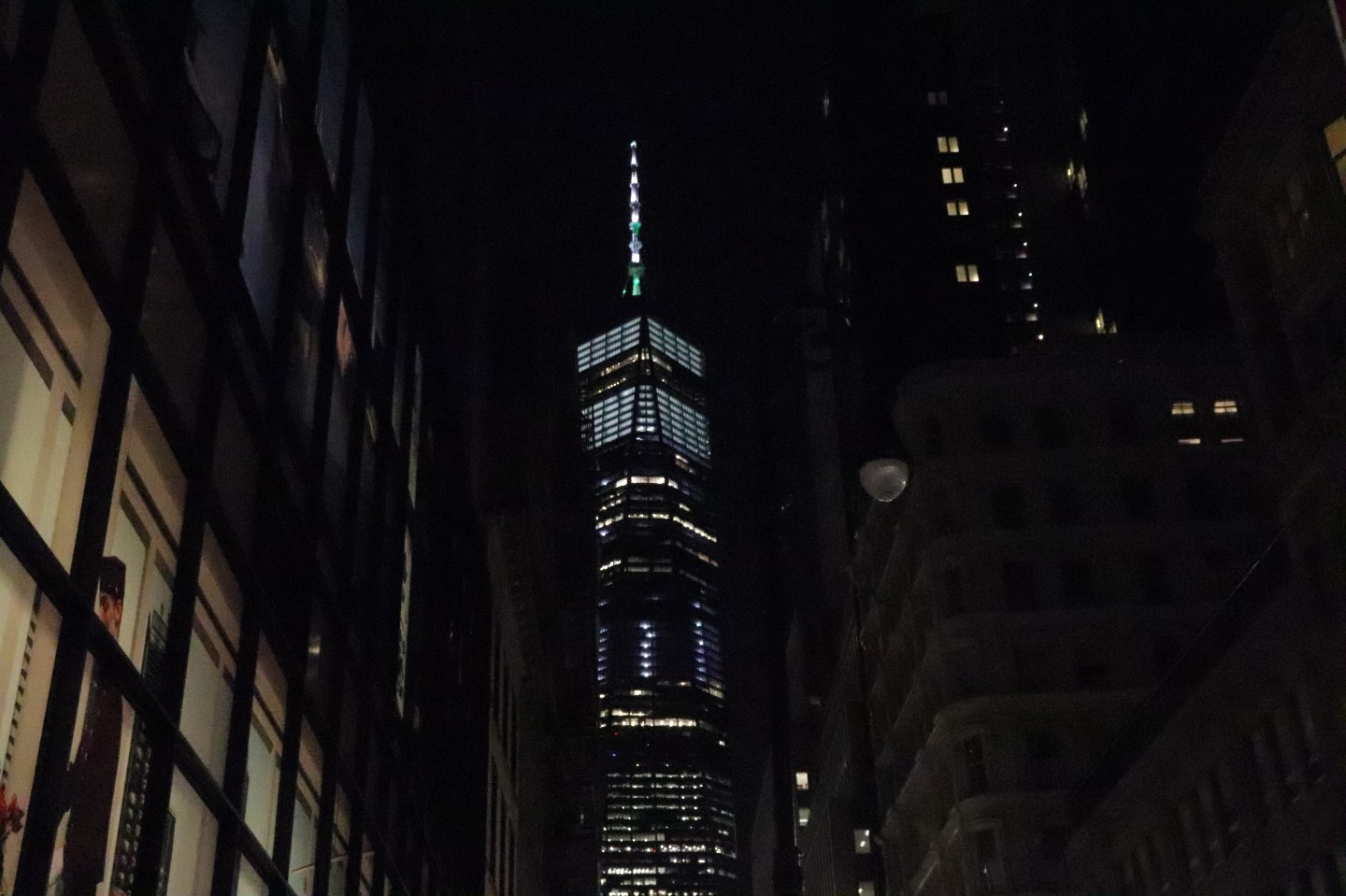 One World Trade Center illuminated between dark buildings at night.